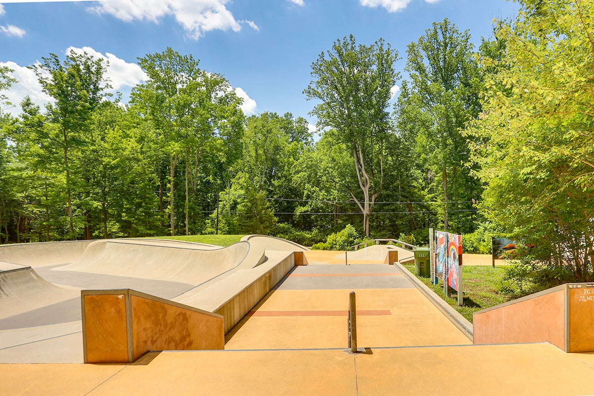 View down a short flight of stairs into a skate park with a smooth skating area, metal railings, and a large concrete bowl on the left.