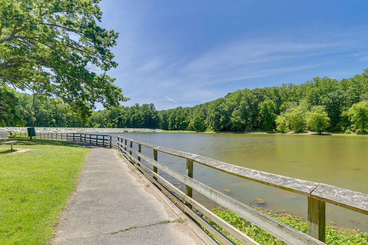 A sidewalk and wooden fence run along the bank of a large, calm body of water. There are trees on the opposite shore.