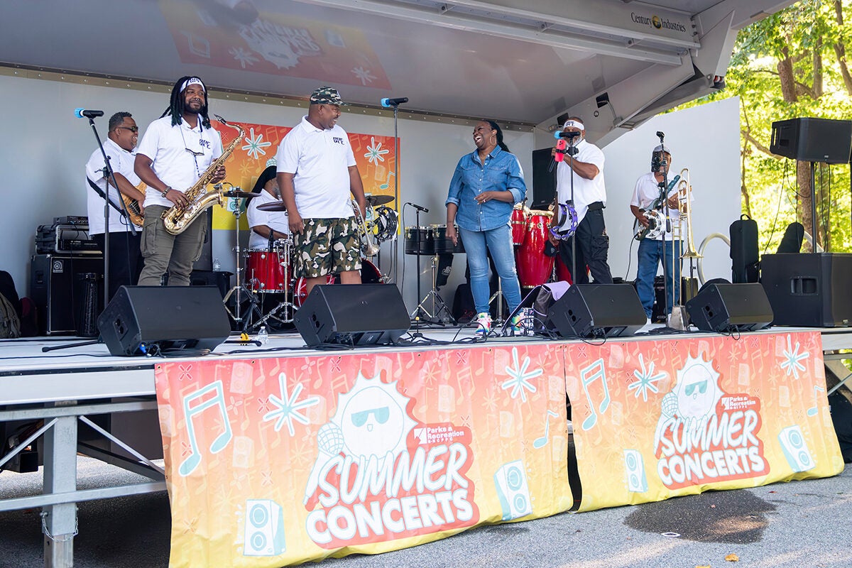A band performs in a mobile bandshell in a park. Two colorful banners attached to the front of the stage read, "Summer Concerts."