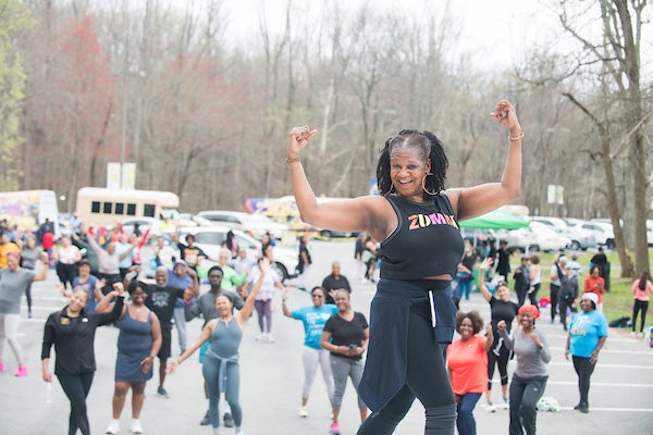 A woman with a shirt that says "Zumba" exercising and demonstrating for a crowd of people behind her.