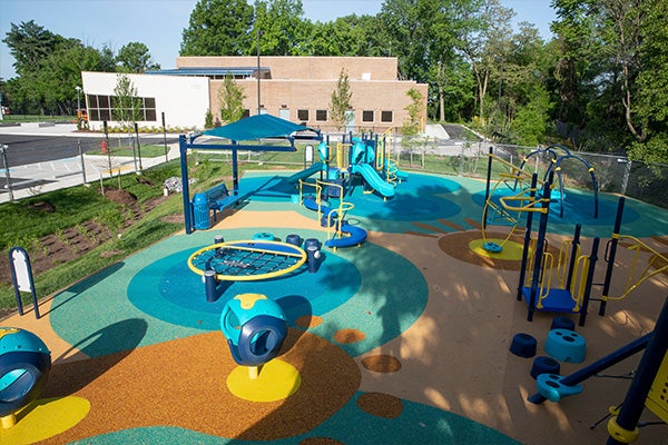 Bird's-eye view of a colorful and shady playground with the brick community center in the background.