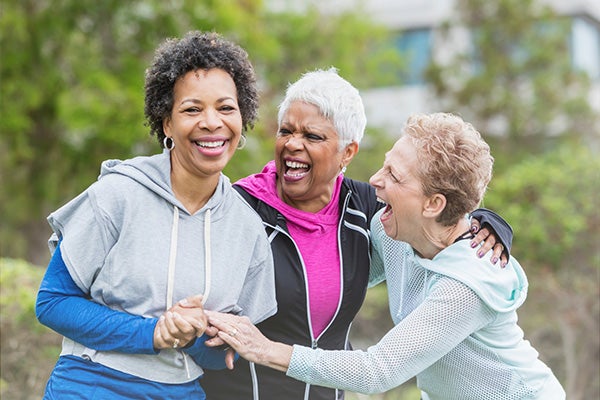 Three senior women outdoors, laughing and clasping hands in an embrace.