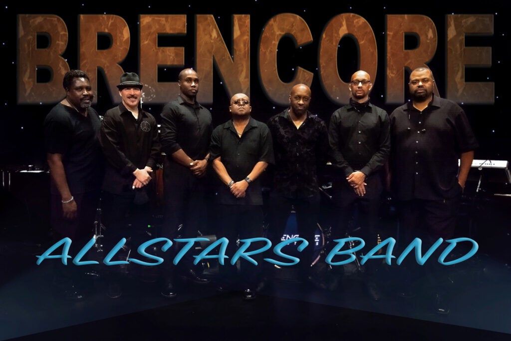 Seven musicians dressed in black stand in a row on stage with band name text behind them.