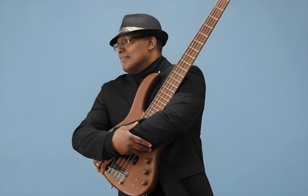Person wearing a hat and jacket holds a bass guitar against a solid blue studio background.