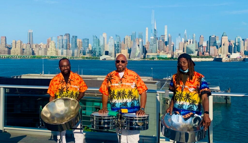 Three musicians wearing orange, yellow, and blue tropical shirts stand with steel drums on a waterfront overlook, city skyline and blue water in background.