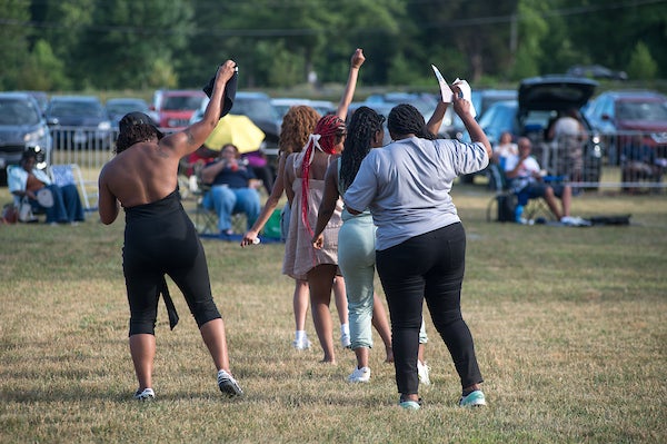 People dancing across a grassy field during an outdoor community event, with parked cars and spectators in the background.