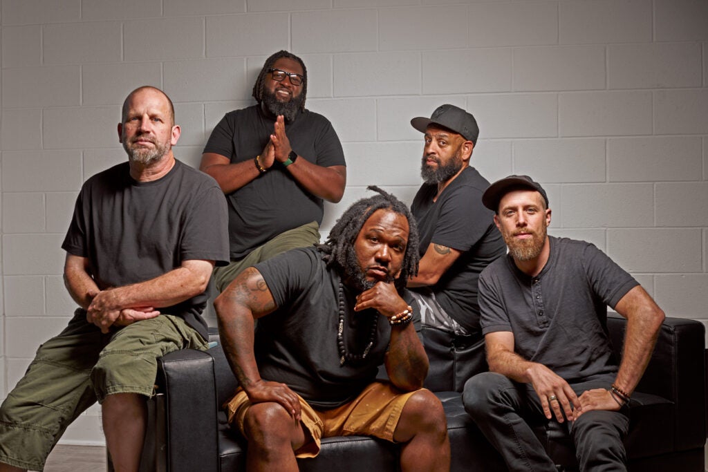 Six people pose together on a studio couch against a gray wall, wearing casual clothing and neutral colors.