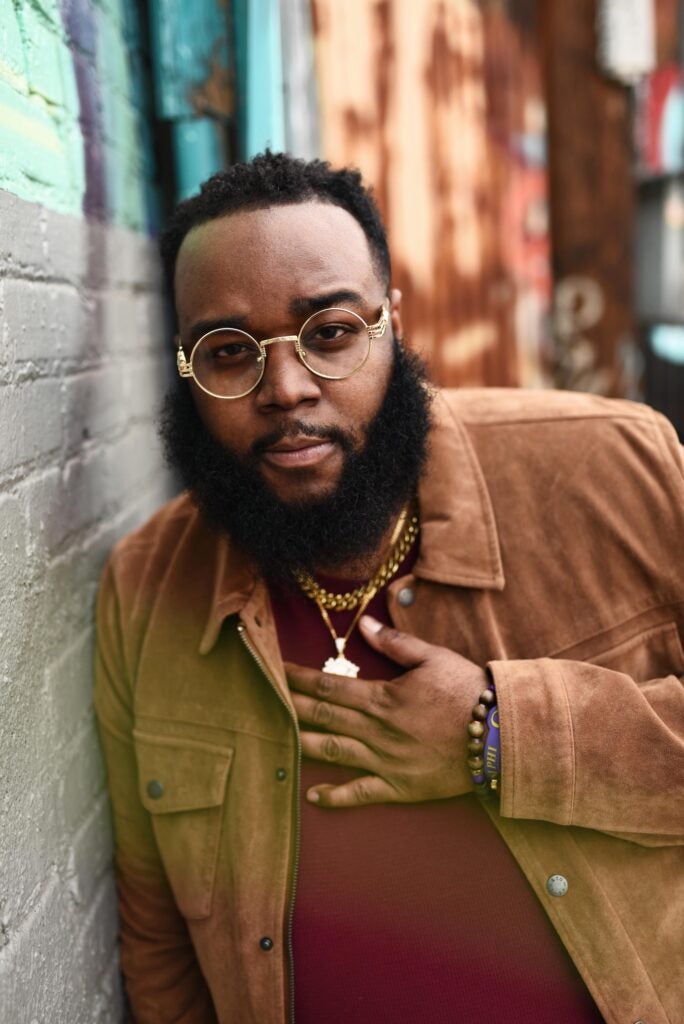 Person wearing glasses and a brown jacket leans against a brick wall in an outdoor urban setting.