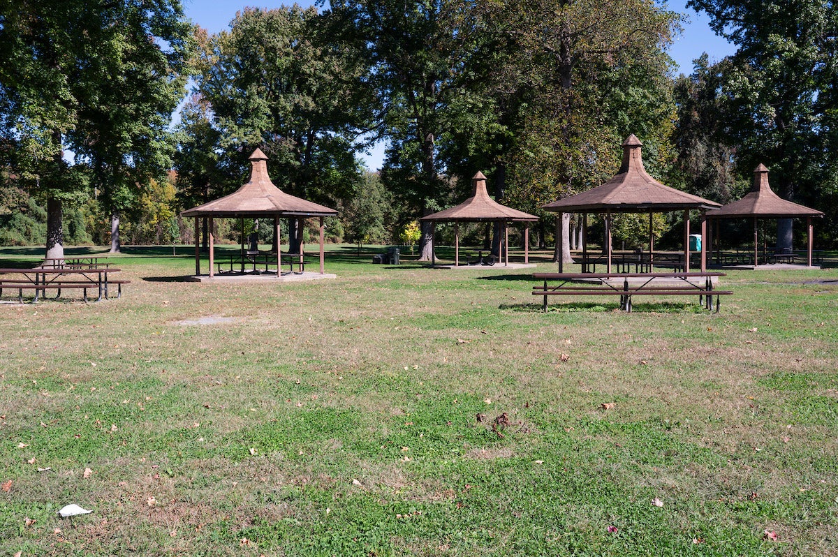 Five small picnic shelters in a field near some trees, each covering one picnic table. There are unsheltered picnic tables nearby.