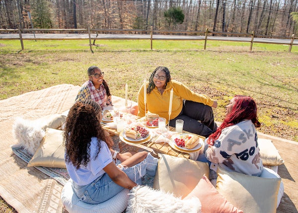 Four people seated on cushions around a low picnic table outdoors, with candles, plates of food, and drinks on layered rugs in a grassy area.