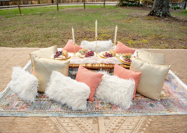 Outdoor picnic on patterned rugs with a low table, candles, plates of food, and cream, blush, and fluffy white pillows arranged on grass under trees.