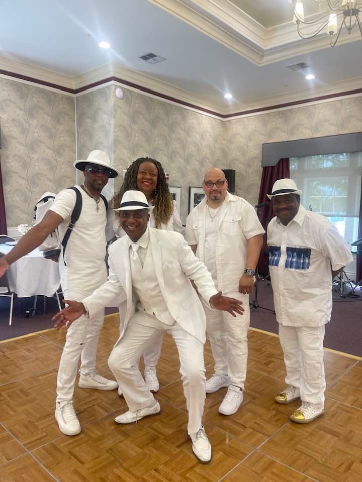 Six people dressed in white pose together on a dance floor inside an event space with tables and decorations.