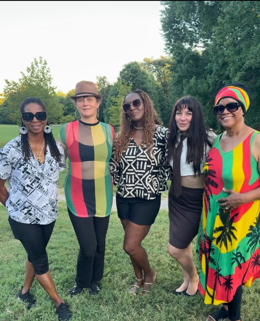 Five people stand together on grass in a park, wearing colorful outfits with trees and open space behind them.