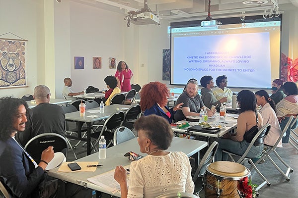 People sit at tables set up in an art gallery while a standing woman presents a slide presentation.