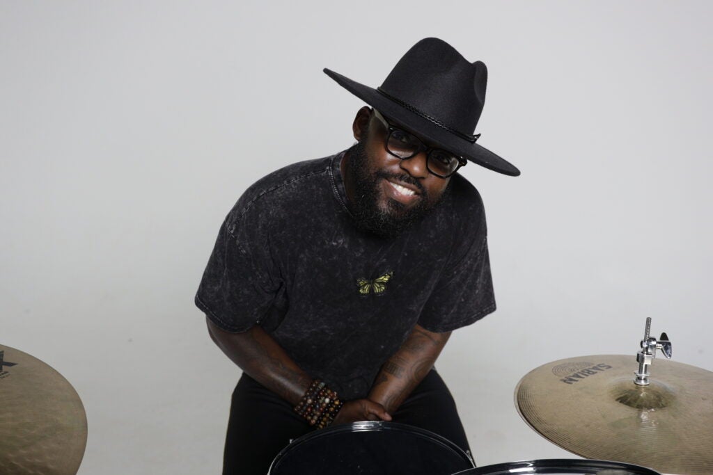 Person in black hat and glasses sits at drum kit, wearing dark shirt and bracelets against plain light background.

