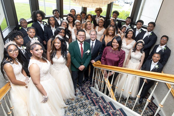 Several dozen people in formalwear pose on the landing of a large stairwell under a chandelier. The young women wear white gowns and tiaras; the young men wear tuxedos.
