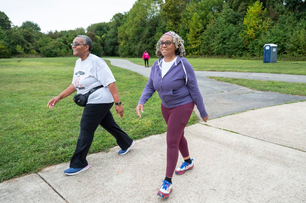 Two people walk on a paved park path with trees behind them, and another person walking in background.
