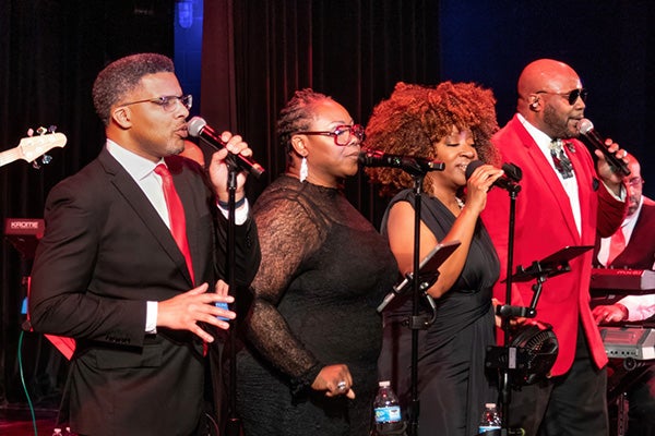 Four adults in formalwear sing into microphones on a stage, with a backing band behind them.