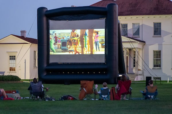 Outdoor movie night with large inflatable screen on lawn; audience seated in folding chairs watching animated film near building.