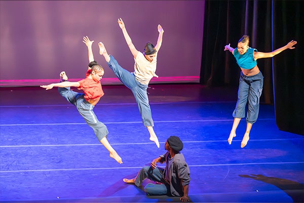 Four dancers perform on a stage. Three are leaping in midair, while one sits on the stage.