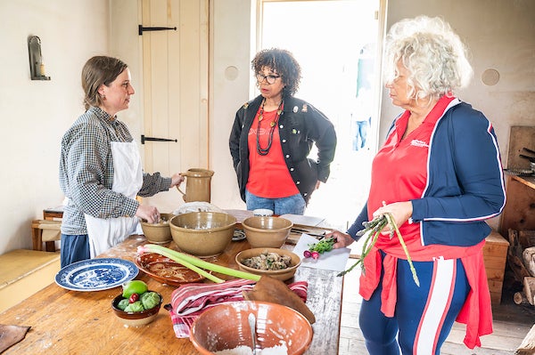 In an 18th-century kitchen, a staff member in a historical costume talks to two visitors while preparing ingredients in crockery bowls at a table.