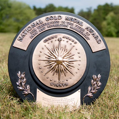A circular, black award in the grass, with metal plates that read National Gold Medal Award for Excellence in the Field of Park and Recreation Management, and Grand Award Winner.