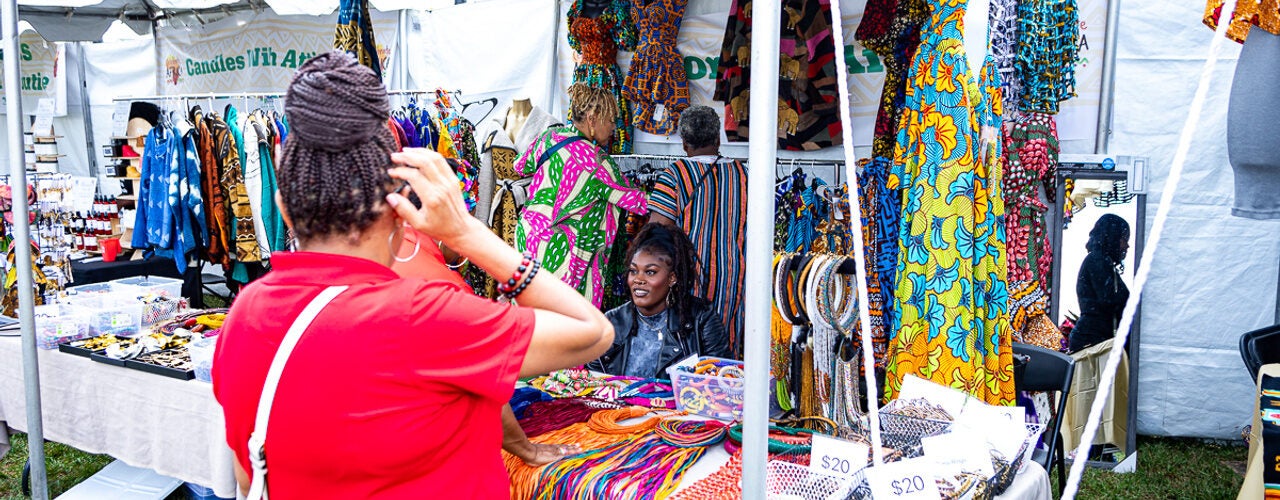 Outdoor festival vendor booth displaying colorful patterned clothing, jewelry, and accessories as a visitor browses merchandise under a white canopy.