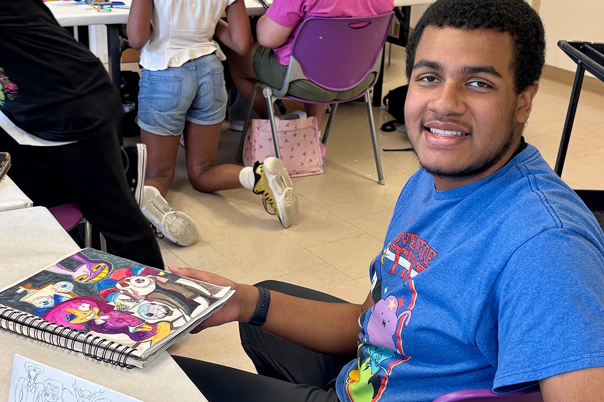 A young person sitting in a classroom holds a sketchbook containing a colorful drawing.