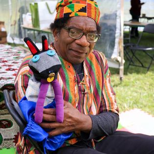 Person in a colorful striped outfit and patterned cap holding a handcrafted puppet beneath a festival tent.