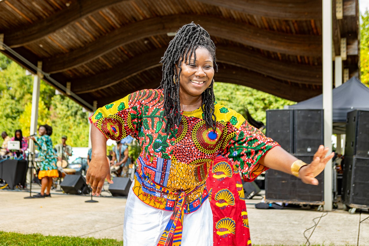 Performer in a brightly patterned outfit dances near an outdoor stage, with musicians, speakers, and festival attendees visible in the background.