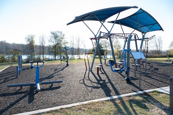 Wide view of an outdoor fitness area featuring multiple exercise stations, shade canopies, and a lake in the background.