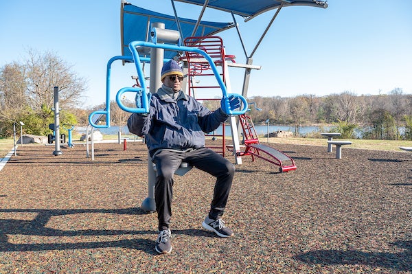 Person seated on an outdoor chest press machine beneath a shade canopy at a lakeside fitness park.
