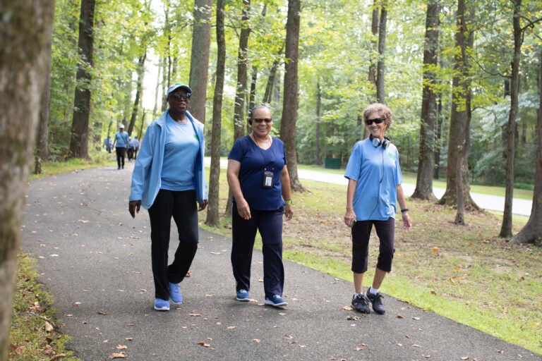Three smiling older women wearing athletic clothing walking together on a paved path through a wooded park.