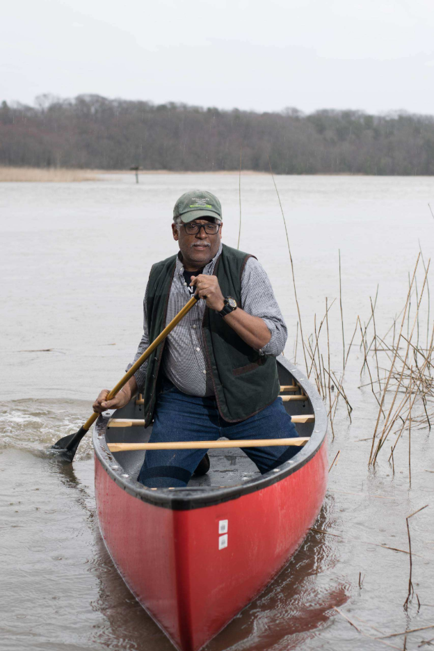 Person paddles a red canoe through shallow water near marsh reeds on an overcast day.
