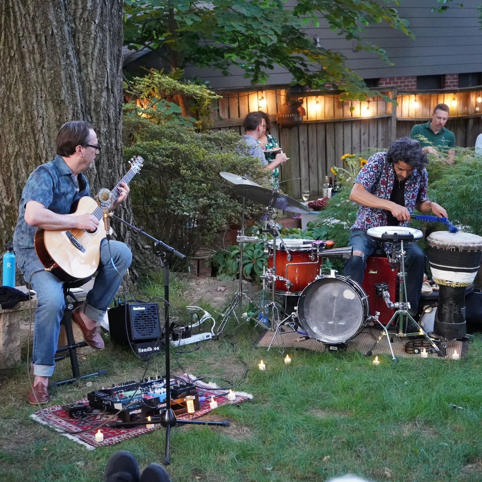 Musicians perform an outdoor garden concert, with an acoustic guitarist and percussionist playing among trees, candles, and seated guests.