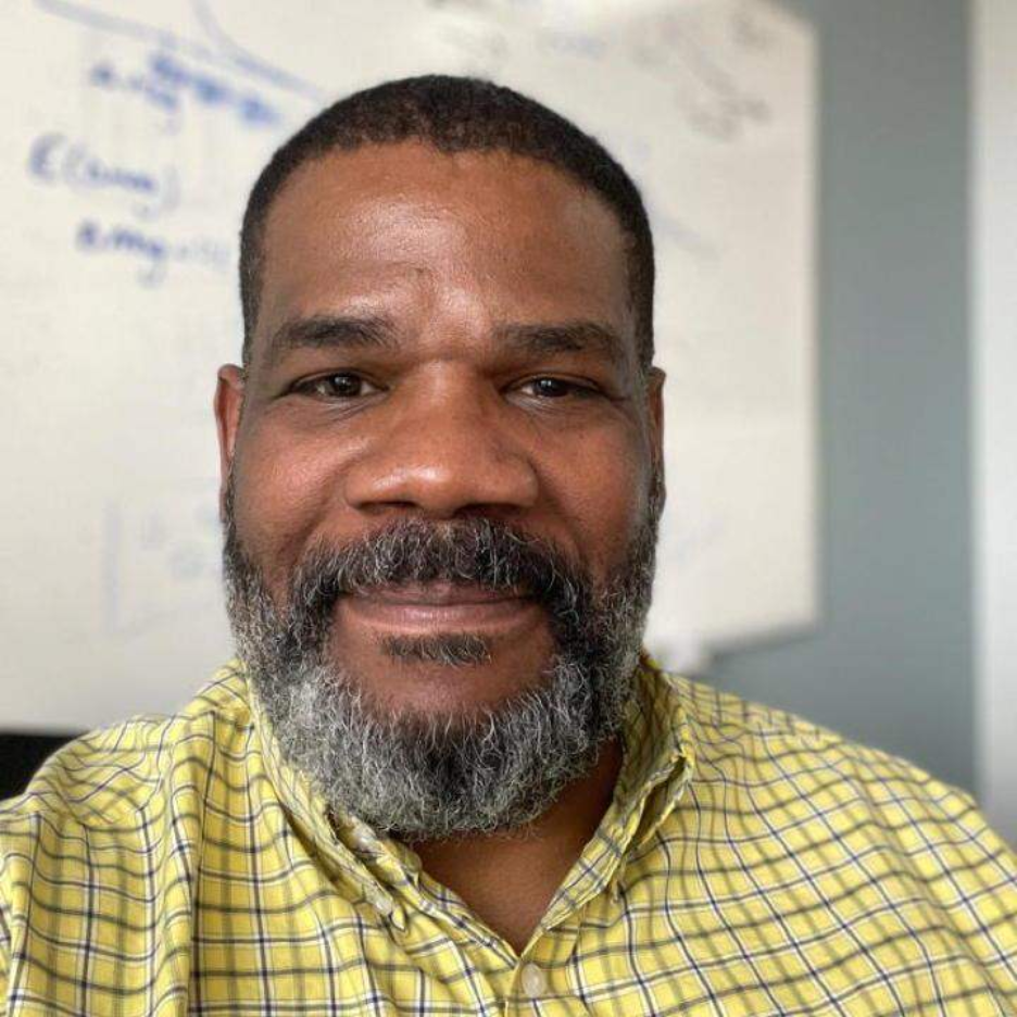 Head-and-shoulders portrait of a bearded person in a yellow plaid shirt seated in front of a whiteboard.