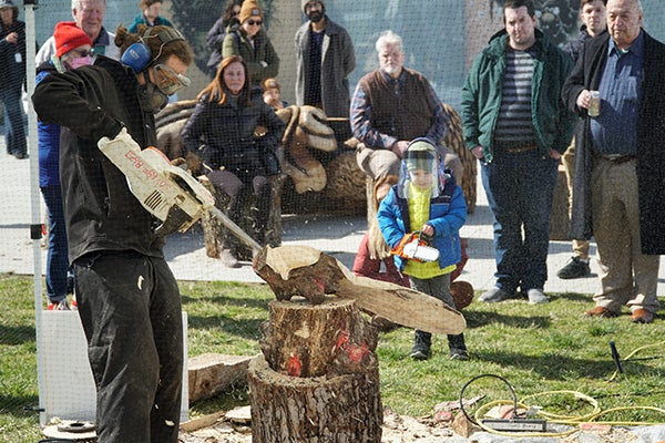 A person in protective gear carves away at wood using a chainsaw. People are gathered around to watch them work.