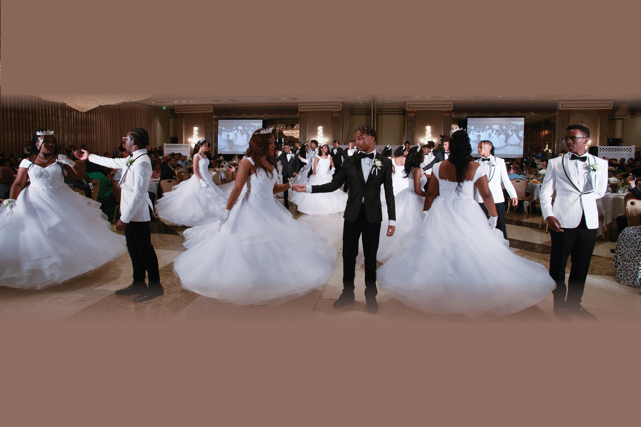Teens perform a choreographed dance, dressed in tuxedos and white dresses. They're dancing in a tan ballroom with displays and large chandeliers in the background.